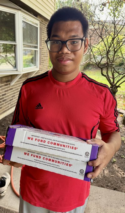 A young man wearing glasses and a red Adidas shirt stands outdoors holding two stacked 'We Fund Communities' ice cream novelty school bus kits.