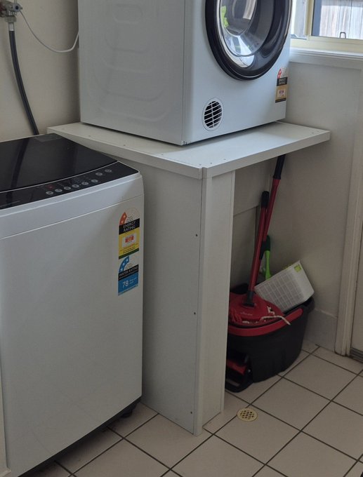 A laundry room setup with a white front-loading washing machine on the floor and another white appliance elevated on a custom platform, with a red mop bucket and cleaning supplies stored underneath.