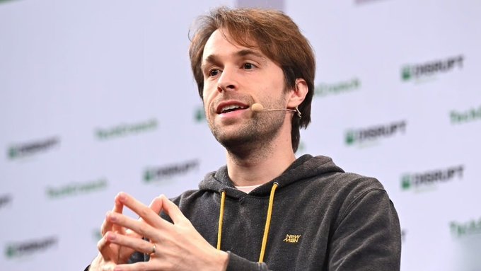 A man wearing a dark hoodie with yellow drawstrings speaks at what appears to be a TechCrunch Disrupt event, gesturing with his hands while wearing a headset microphone.
