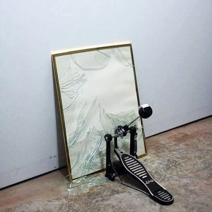 A broken floor mirror leaning against a wall, with a Ludwig bass drum pedal lying on top of the shattered glass on the floor.