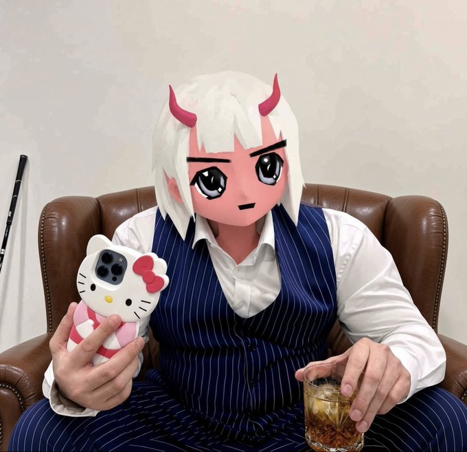 A person with a cartoonish anime-style virtual filter (white hair and red horns) wearing a navy pinstripe vest sits on a leather couch, holding a Hello Kitty phone case and a glass of tea.