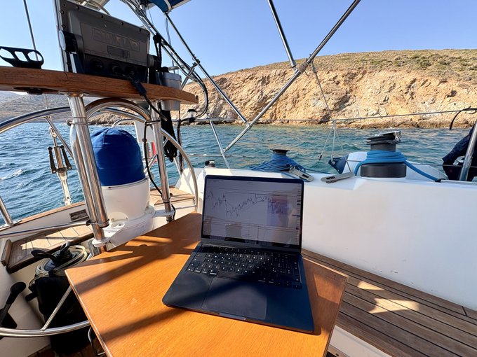 A laptop sits open on a wooden table aboard a sailboat, displaying a chart, with rocky cliffs and blue water visible in the background.