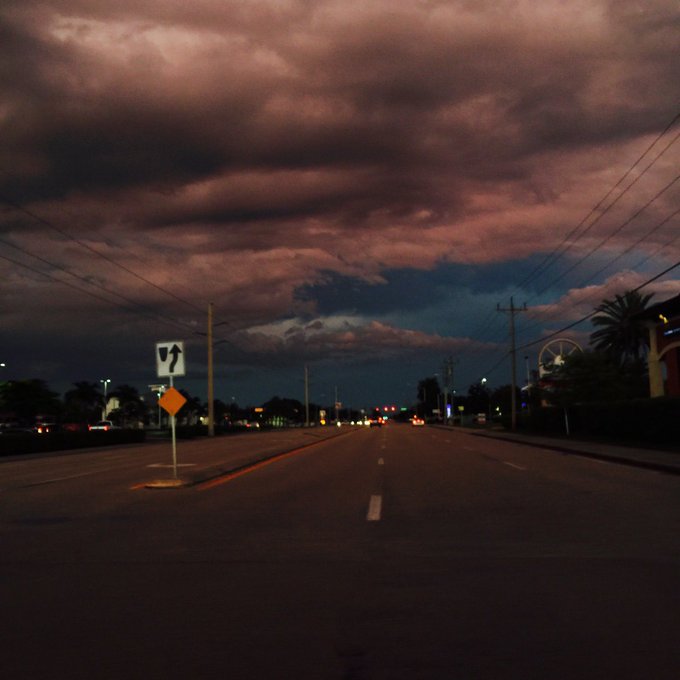 A nighttime view down an empty road with dramatic purple-pink clouds in the sky, a divided highway sign on the left, and a Ferris wheel visible in the distance on the right.