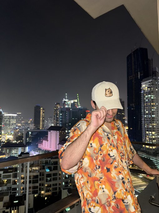 A man wearing an orange shirt covered in Shiba Inu (Doge) faces stands on a rooftop at night with a city skyline behind him, adjusting his white cap which features a small Shiba Inu logo. The image illustrates the post's playful claim that his Doge-themed shirt outshines Andrew Tate's, while promoting $Doge over "Casino" tokens.