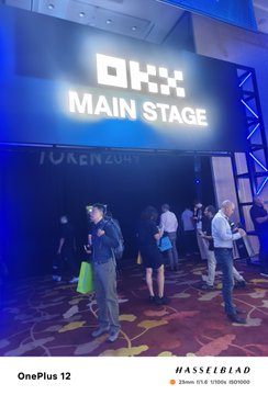 A crowd gathered near a stage area with a large illuminated sign reading 'OKX MAIN STAGE', with watermark text 'OnePlus 12' and 'HASSELBLAD' visible.