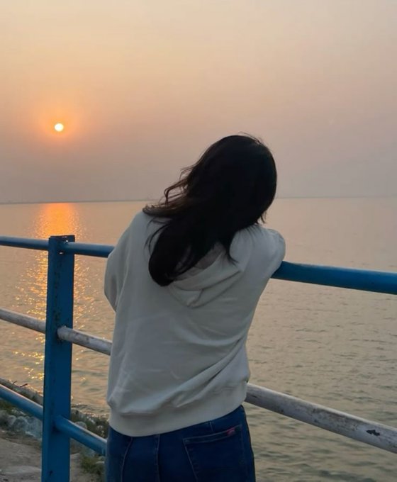 A person with long dark hair, wearing a light grey hoodie and blue jeans, stands at a blue railing looking out at a sunset over the ocean.