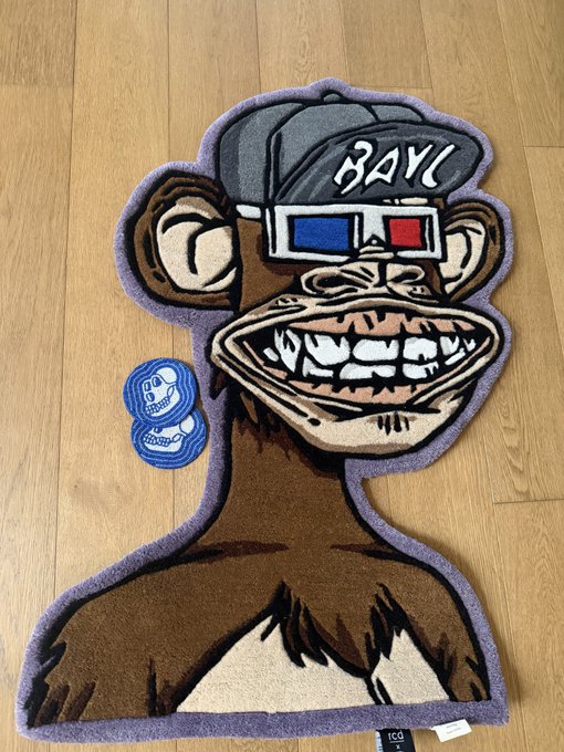 The image features a monkey rug with a cartoon character design, including a hat and glasses, placed on a wooden floor. The rug has a purple border around its edges, giving it an artistic touch. There are two stickers attached to the bottom of the rug, one blue and one white. A caption below the image reads: "Never been more comfortable getting rugged !!! Thanks to @richclasdecor for the efficiency of this transaction".