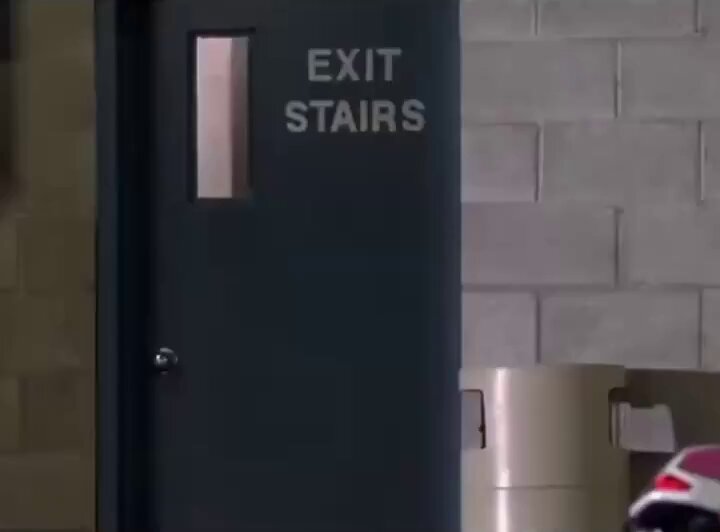 The image shows a black door with white text, which reads "EXIT STAIRS", located inside an office building. The door has a window above it, allowing some light into the room. In front of the door, there's a red motorcycle parked against the wall. A person is also visible in the background, standing near the motorcycle and the door.