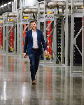A man in a blue suit walks down an aisle of a large data center, with server racks labeled A053, A054, and A056 visible behind him. The image likely depicts a facility related to American Bitcoin's mining operations, contextualizing the article about the company's dramatic stock decline and Eric Trump's significant financial losses.