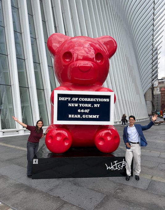 Two people posing on either side of a large red gummy bear sculpture at what appears to be the World Trade Center Oculus in New York. The bear holds a sign reading 'DEPT. OF CORRECTIONS, NEW YORK, NY, 6-6-07, BEAR, GUMMY' with the 'WHISTLE' artist signature on the base.
