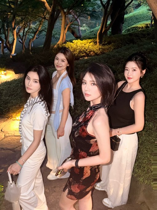 Four stylish women pose together at an elegant outdoor evening event, surrounded by lush greenery and warm landscape lighting that creates a sophisticated garden-party atmosphere fitting for the upscale BNB Chain gathering.