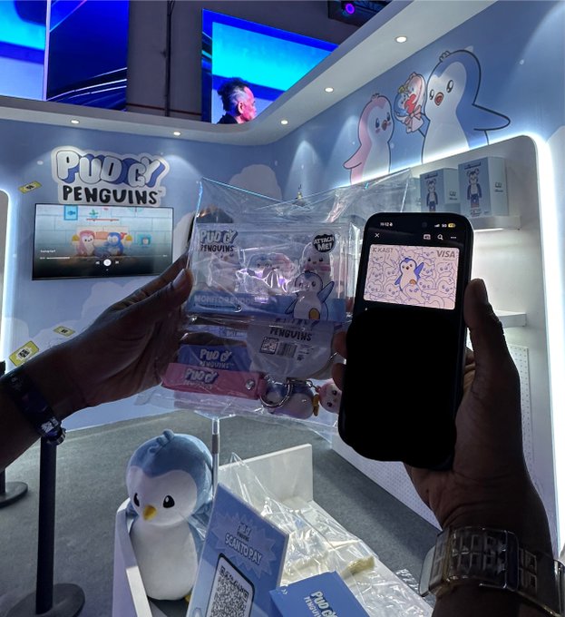 A person holds up a Pudgy Penguins merchandise package and a phone displaying a digital Pudgy Penguins Visa card at a Pudgy Penguins branded booth.