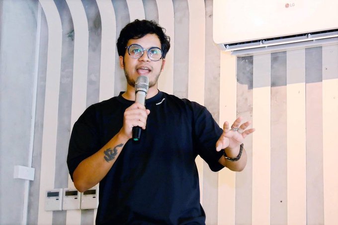 A man with glasses and a black t-shirt speaks into a microphone while gesturing with his left hand.