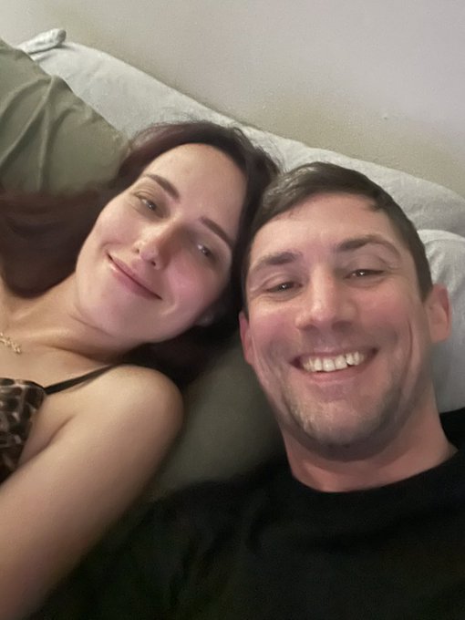 A couple smiles in a close-up selfie while lying together on a bed with light-colored pillows.