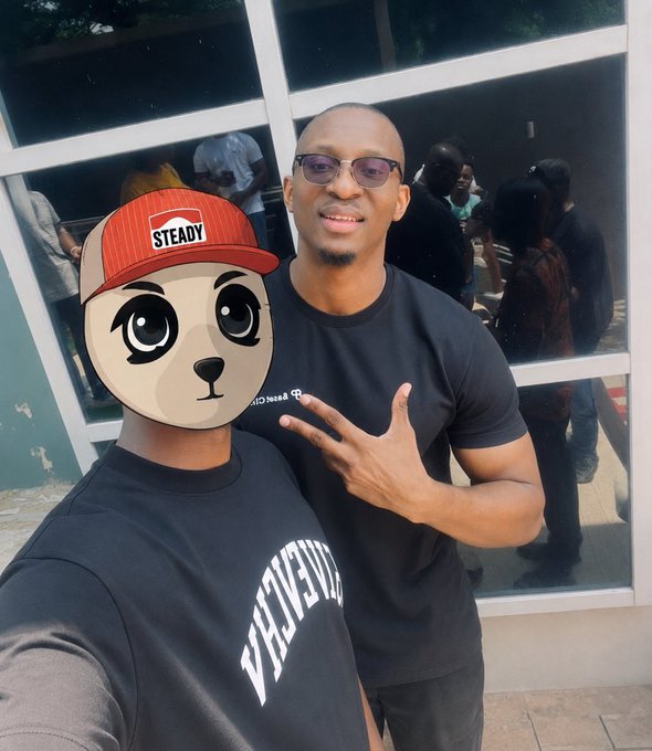 Outdoor photo of two men; one wearing a black "UNIVERSE" shirt has his face covered by a cartoon dog sticker wearing a red "STEADY" cap, while the other wears sunglasses, a black t-shirt, and makes a peace sign near a building entrance.