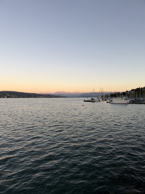 A serene sunset view over a body of water with several boats moored in the distance and a half moon visible in the sky.