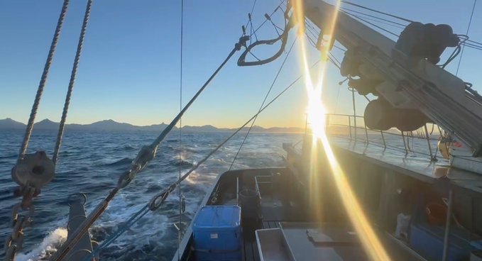 A view from the deck of a boat sailing on the ocean at sunset, with bright sun glare, rigging, and distant mountains visible on the horizon.