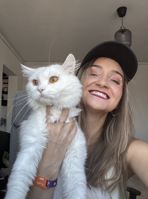 A smiling young woman wearing a black cap holding up a fluffy white cat with yellow eyes, taking a selfie indoors near a window.
