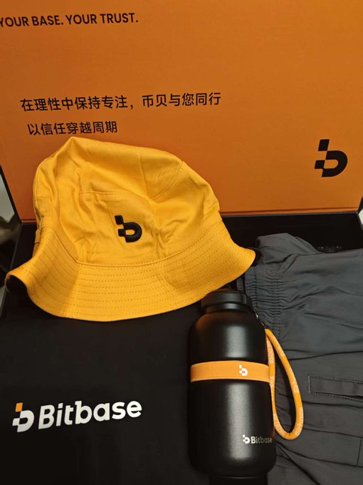 A promotional product display from Bitbase featuring a yellow bucket hat with the 'b' logo, a black water bottle with an orange strap, and gray clothing, all branded with the Bitbase name and logo.