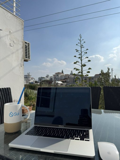 An outdoor workspace setup with a MacBook Air displaying code, a white 'Sui' branded mug, a wireless mouse, and a glass table overlooking a town with an agave plant in the background.