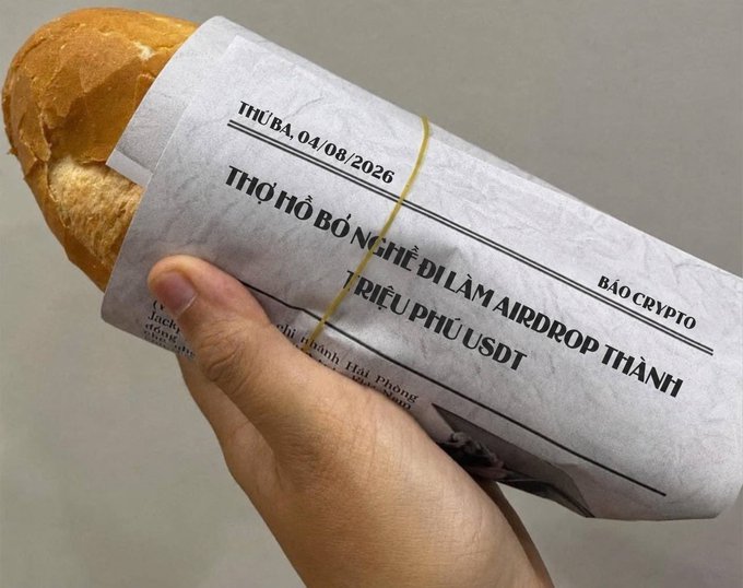 A hand holding a baguette wrapped in paper that has been printed to look like a newspaper with Vietnamese text, including 'THỦ BA, 04/08/2026', 'THƠ TÌO BỘ NGHE DỊ LÀM AIRDROP THÀNH TRIỆU PHÚ USDT', and 'BÁO CRYPTO', a humorous commentary on crypto news.