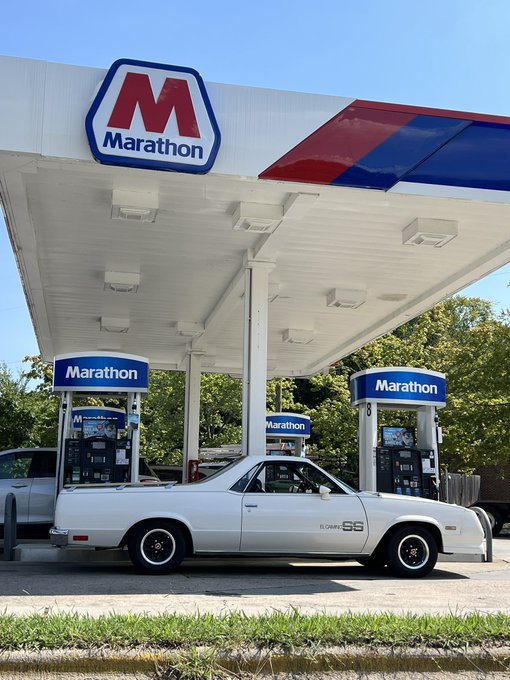 A white classic El Camino SS is parked at a Marathon gas station, with the iconic red and blue Marathon logo prominently displayed on the canopy above the fuel pumps.