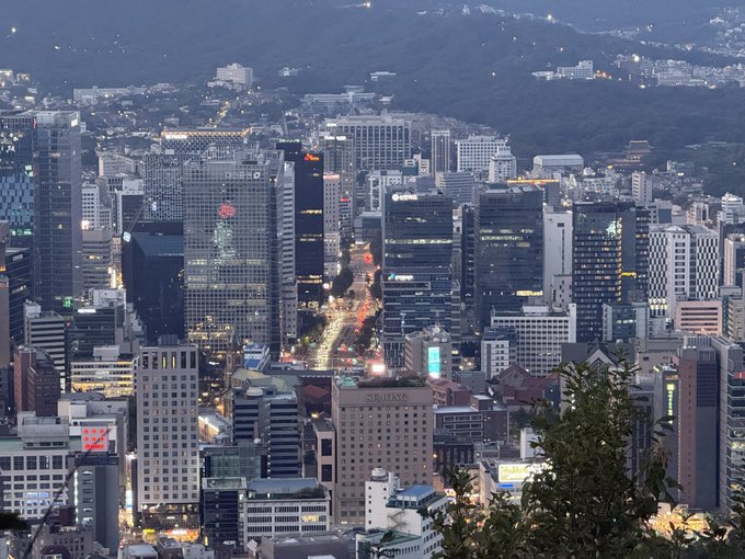 A dusk cityscape of Seoul's dense downtown skyscrapers lights up as night falls, with the "SEJONG" building visible among the towers, matching the post about attending the XRP Seoul crypto meetup in South Korea.