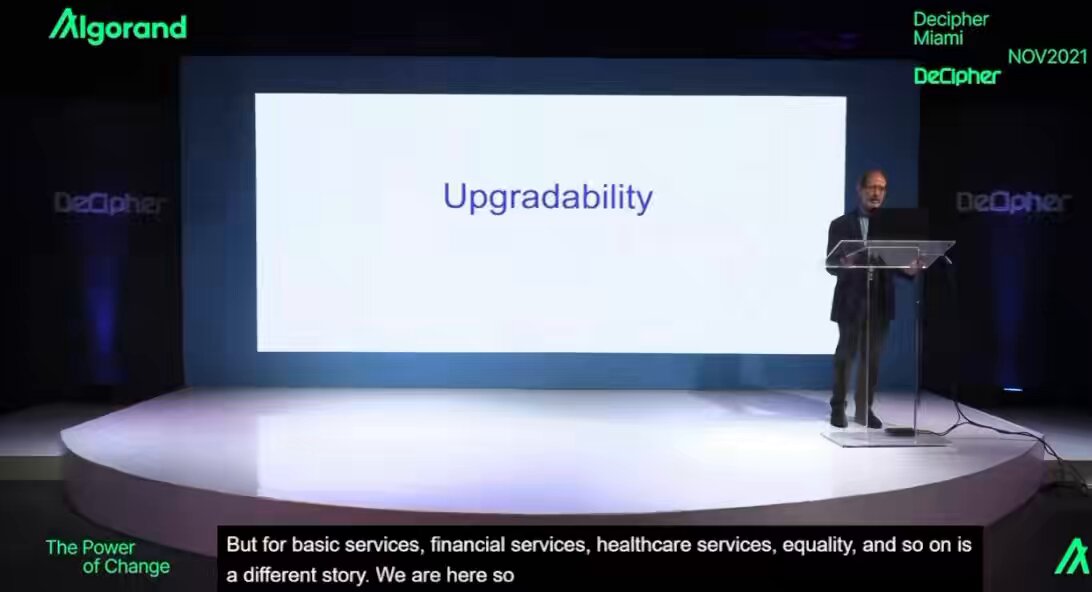 The image shows a man standing at a podium, giving a presentation or speech. He appears to be talking about upgrading a system or service, possibly related to Algorand. The background features a large screen displaying the text "Upgrading" on it.