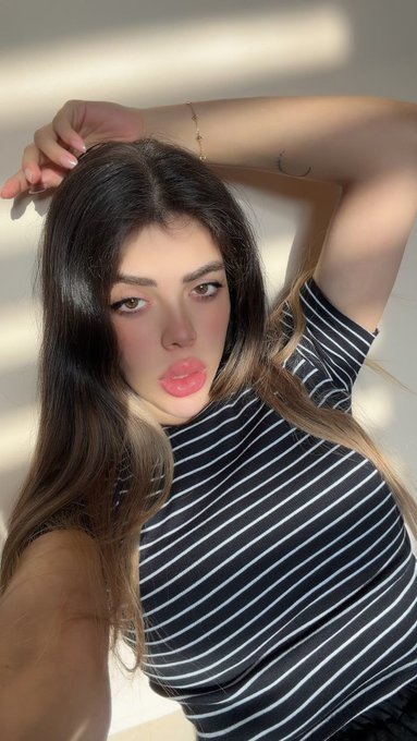 A young woman with long brown hair wearing a black and white striped top poses with her arm raised above her head, sunlight casting shadows across her face.