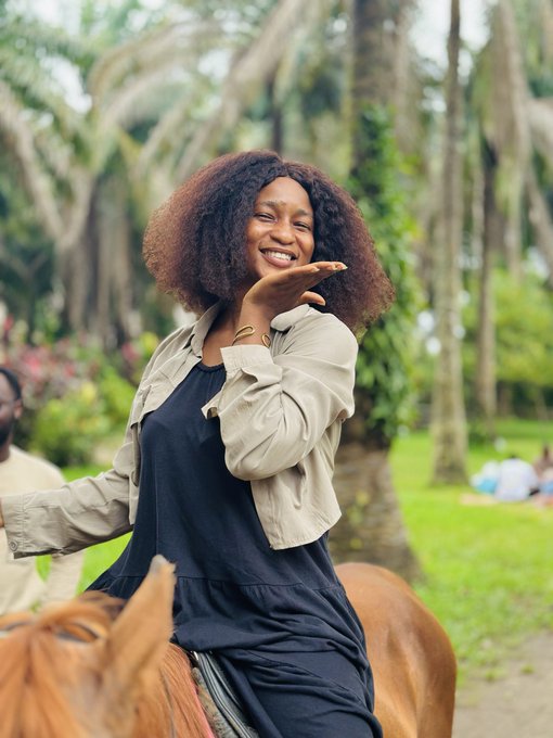 A smiling woman with curly hair rides a brown horse through a lush, tropical setting with palm trees, wearing a black dress and a light jacket.