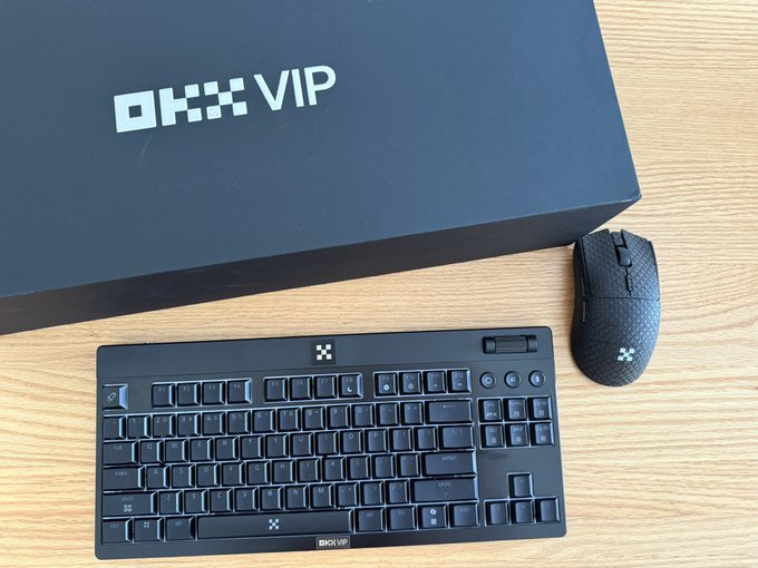 A black OKX VIP box sits atop a wooden desk next to a black OKX-branded keyboard and gaming mouse, showcasing the premium peripherals gifted by the cryptocurrency exchange.