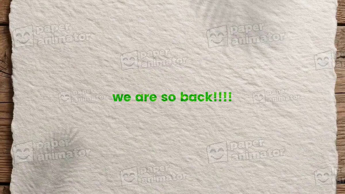 The image features a white paper with green text that says "we are so back!" written in bold letters, placed on a wooden surface. The background appears to be a gray wall or a wooden table. There is no visible action taking place in the image, and there are no other objects present besides the paper and the wooden surface.