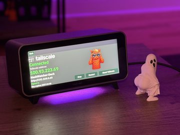 A small black touchscreen device on a desk displays a Tailscale connection status showing an IP of 100.93.223.69, accompanied by a small white ghost figurine and an orange logo icon.