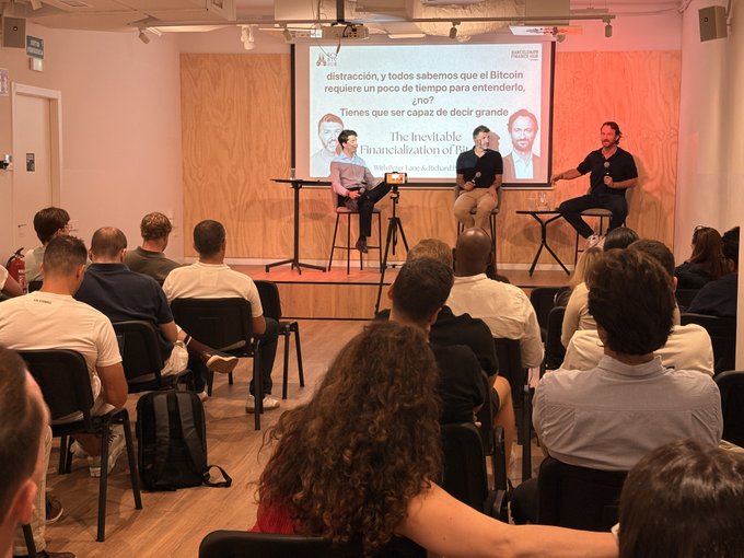 A panel discussion titled "The Inevitable Financialization of Bitcoin" with subtitles about Bitcoin requiring time to understand, featuring three men seated on stage addressing an engaged audience at what appears to be a Barcelona Bitcoin Hub event.
