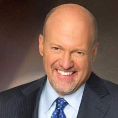 A professional headshot of a bald man with a goatee wearing a dark suit jacket, light blue shirt, and dark blue patterned tie, smiling against a gray background.