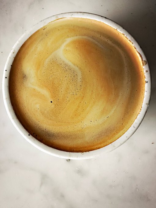 A top-down view of a small white ceramic cup filled with a frothy coffee beverage, likely a cortado or piccolo, resting on a marble surface.