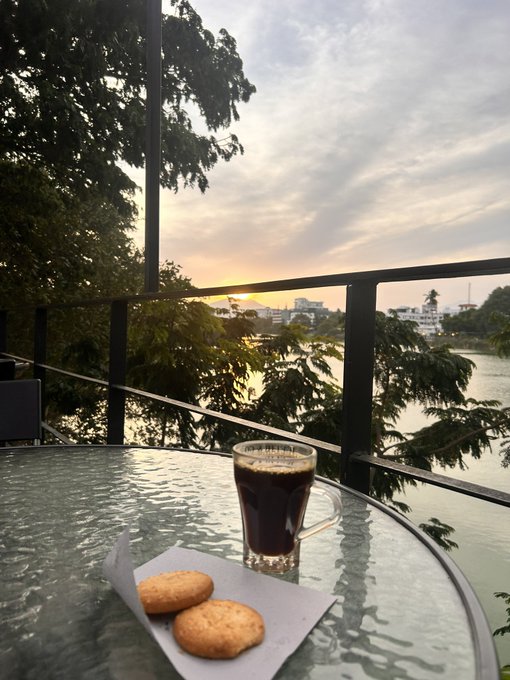 A glass of dark coffee and two cookies sit on a glass table overlooking a scenic river at sunset, with trees framing the view.