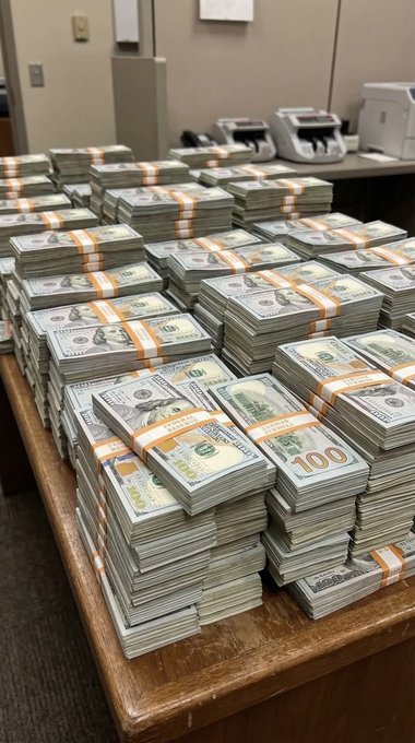 Stacks of bundled U.S. $100 bills are arranged on a wooden table, appearing to be in a money-counting facility with processing machines visible in the background.