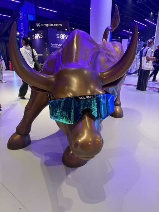 A bronze bull statue wearing iridescent blue "SOLANA" branded goggles is displayed at the Solana BreakPoint conference, with crypto.com signage visible in the background. The life-sized mascot embodies the bullish spirit of the annual Solana ecosystem event that brings together founders, investors, and community members worldwide.
