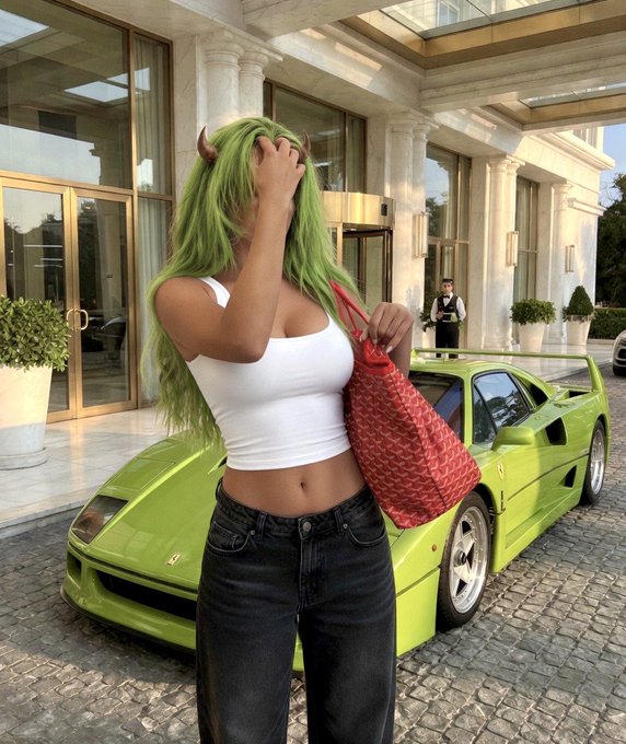 A woman with long green hair wearing a white tank top and dark jeans, holding a red patterned bag in front of a bright green Lamborghini at what appears to be a luxury hotel entrance.