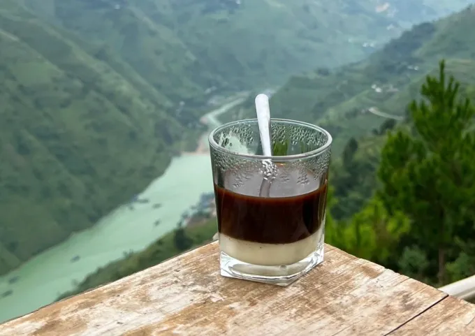 A glass of coffee with a spoon sits on a wooden ledge overlooking a scenic river valley surrounded by green mountains.