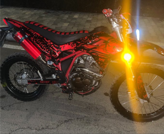 A red and black 'heuer' branded dirt bike with glowing yellow headlights parked at night on a paved surface.