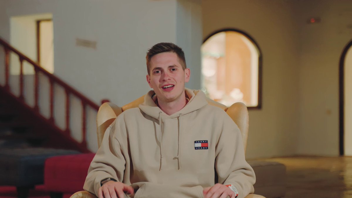 The image features a man sitting in a chair wearing a beige hoodie that has an "Tommy Hilfiger" logo on it. The man appears to be smiling for the camera while seated, possibly posing for a photo or video. The background of the image shows stairs leading up to another room and a door, suggesting that this scene takes place in a house or apartment setting.