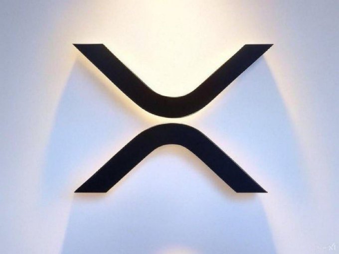 A black, illuminated XRP logo is mounted on a white wall, casting a soft glow around the stylized "X" shape. The image accompanies a post promising $100 to everyone if the XRP cryptocurrency reaches $5.00 within 48 hours, visually tying the promotional claim to the XRP brand.