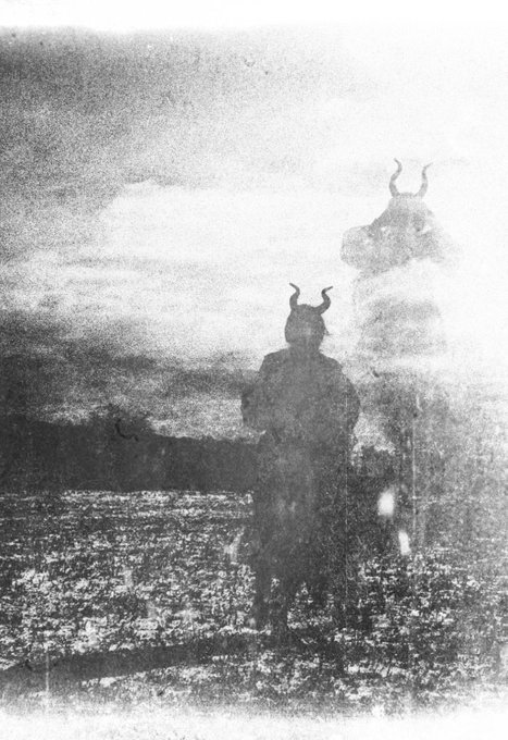 A black-and-white, vintage-style photograph of a solitary figure wearing a horned helmet standing in a desolate landscape, with a ghostly double-exposure silhouette visible behind them.