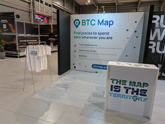 The focus of the image appears to be a booth, possibly at an event or exhibition related to Bitcoin or cryptocurrency. A white sign displaying "THE MAP IS THE MAP" can be seen inside the booth, accompanied by a banner that says "We are ready for you @BTCPrague". The background indicates that this could be taking place in a convention center, as there is a large screen visible behind the booth and other booths can also be seen.