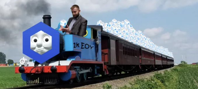 A surreal composite image showing Thomas the Tank Engine's face as a hexagonal logo on the front of a steam locomotive, with a man in a suit holding a phone and a trail of jellybeans billowing as smoke.