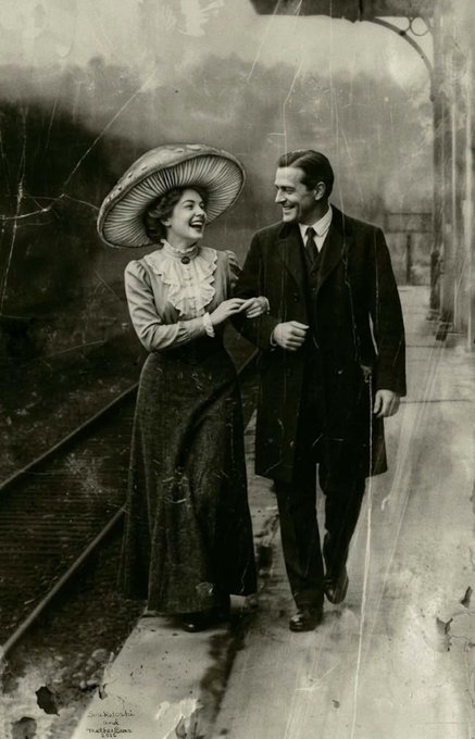 A vintage black-and-white photograph showing a smiling Edwardian-era couple walking arm-in-arm along a railway platform, the woman wearing an oversized hat and ruffled blouse, with handwritten text in the lower left corner.
