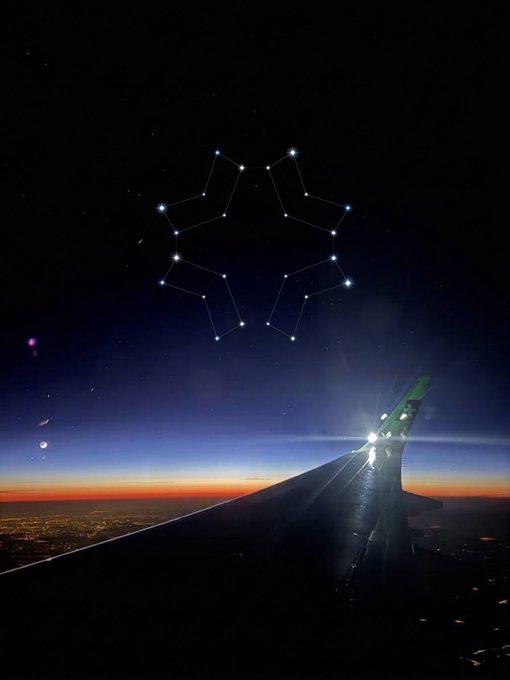 A view from an airplane window at sunset, showing the wing silhouetted against a darkening sky with a starry night above, featuring a constellation pattern resembling a geometric figure.
