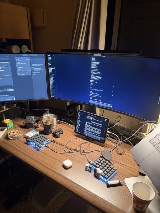 A developer's home workspace at night, featuring a triple-monitor setup with code displayed on two screens alongside a tablet, illuminated by RGB mechanical keyboards, with a coffee cup, Rubik's cube, and snacks scattered across the desk—capturing the humble setup where future success is being built.